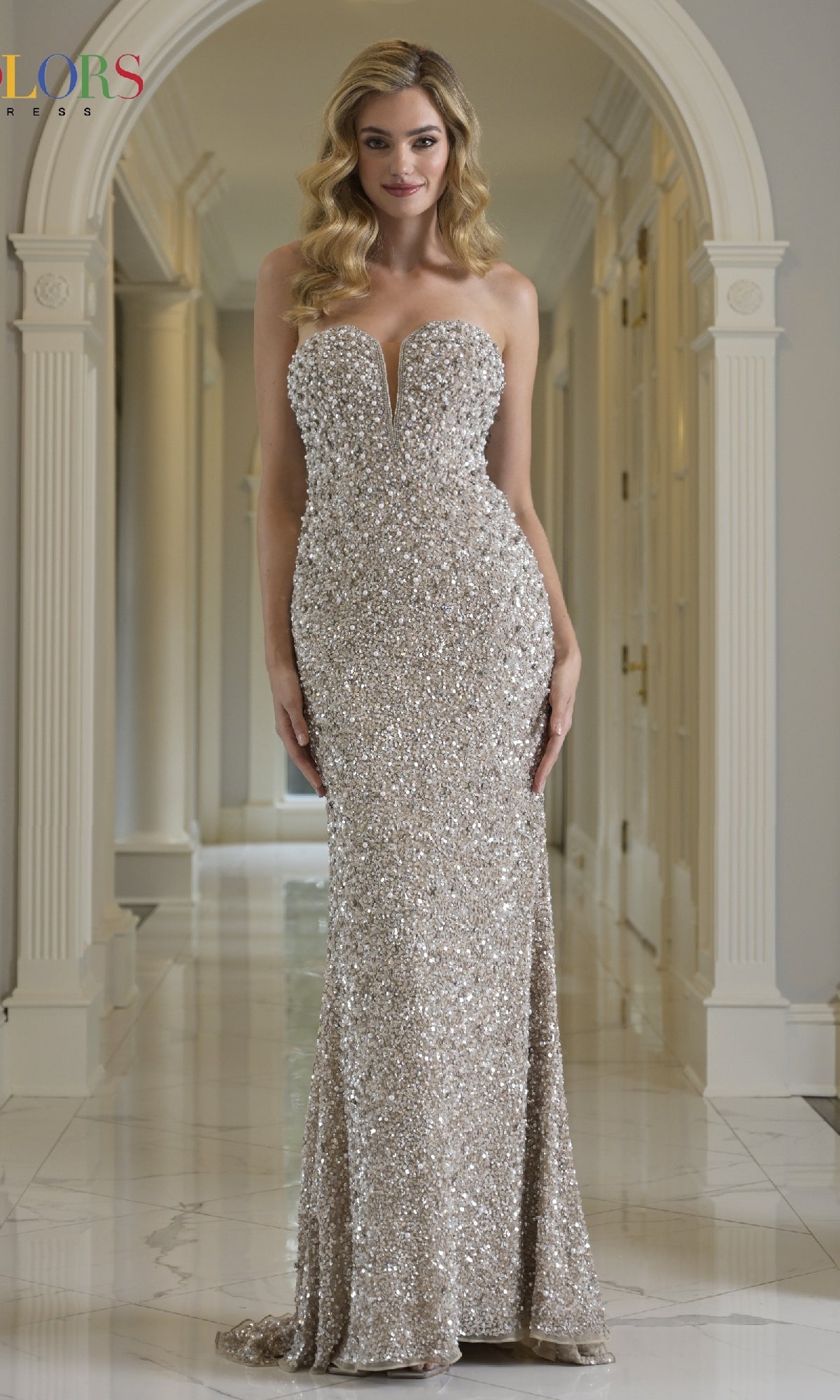 A woman stands in a hallway with arched doorways and marble floors, wearing the shimmering floor-length silver sequin Long Prom Dress: Colors Dress 3939.